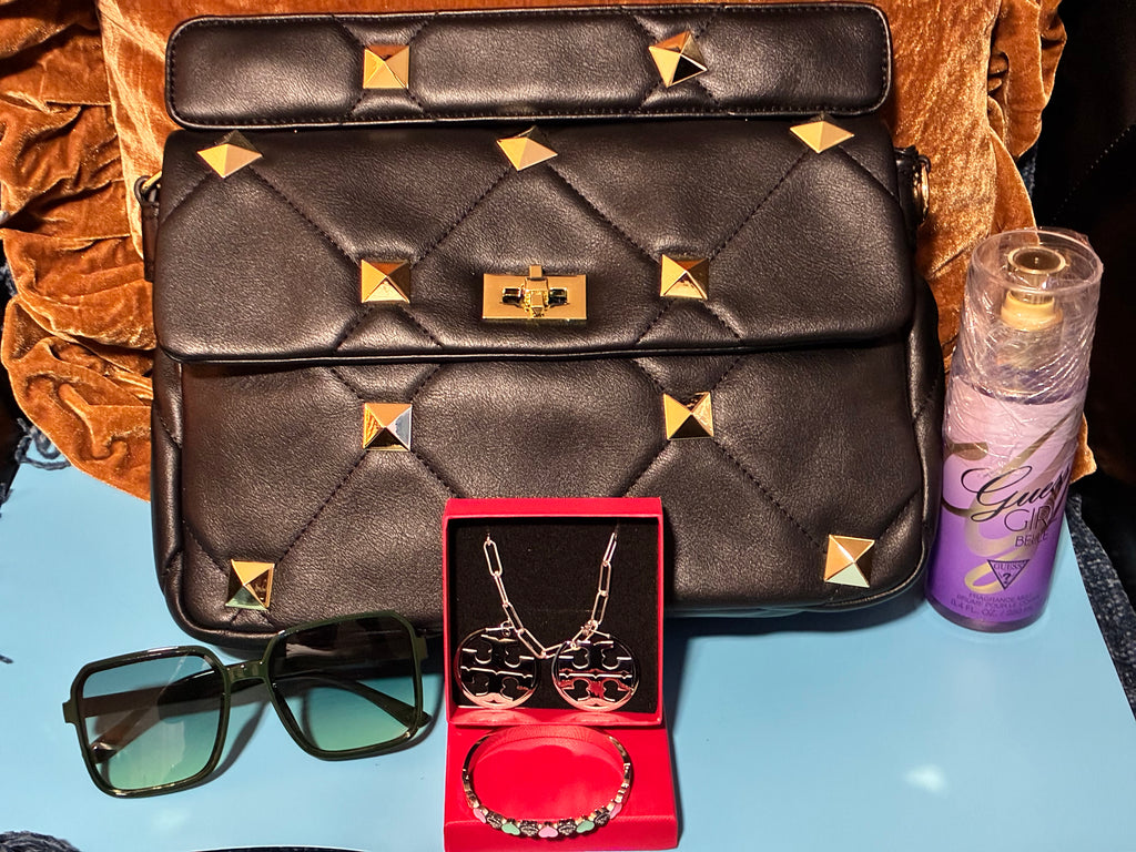 Black quilted handbag with gold studs, sunglasses, jewelry set, and purple bottle on a blue surface.