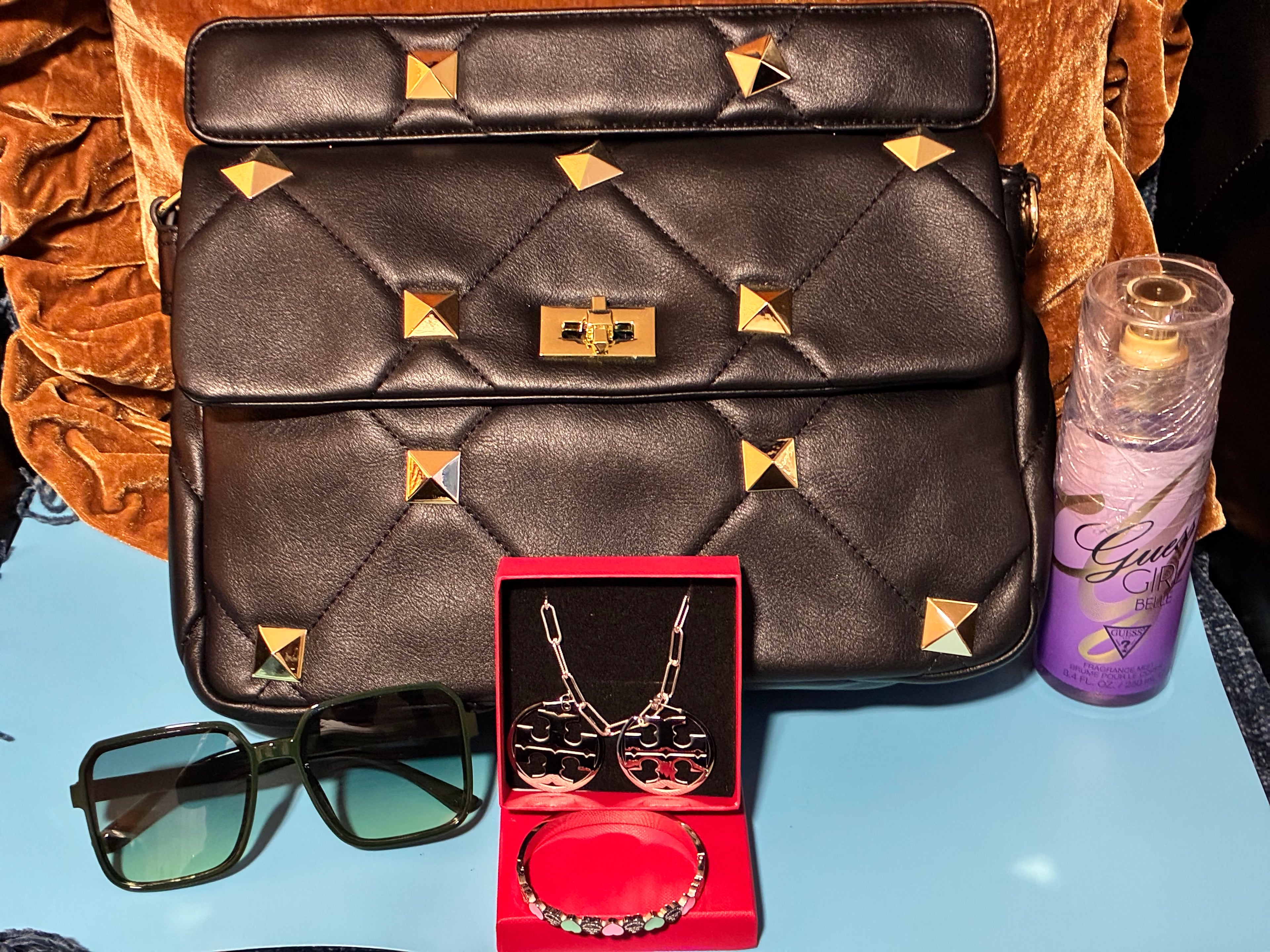 Black quilted handbag with gold studs, sunglasses, jewelry set, and purple bottle on a blue surface.