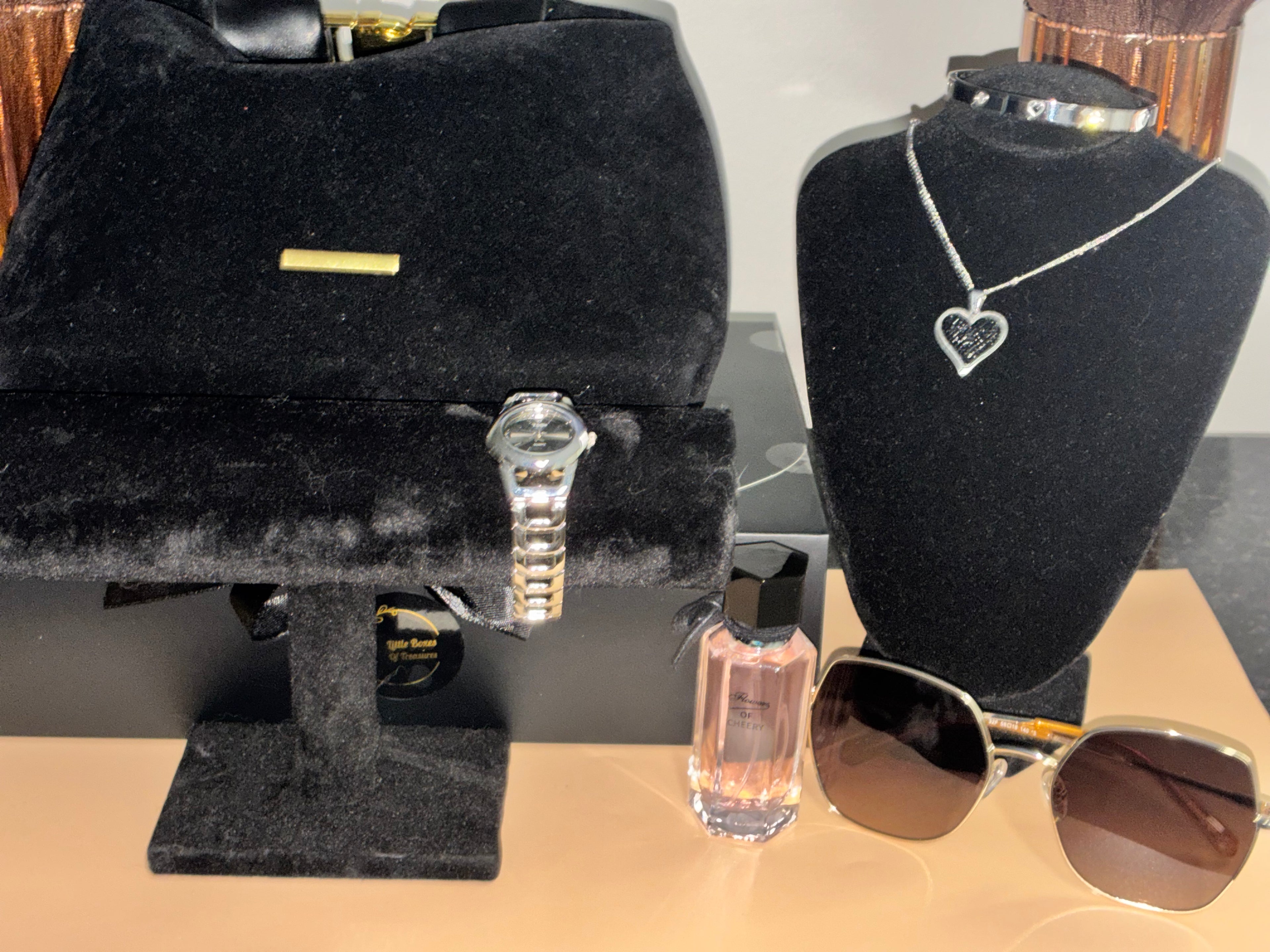 Jewelry display with a black velvet box, silver watch, perfume bottle, and sunglasses on a wooden surface.