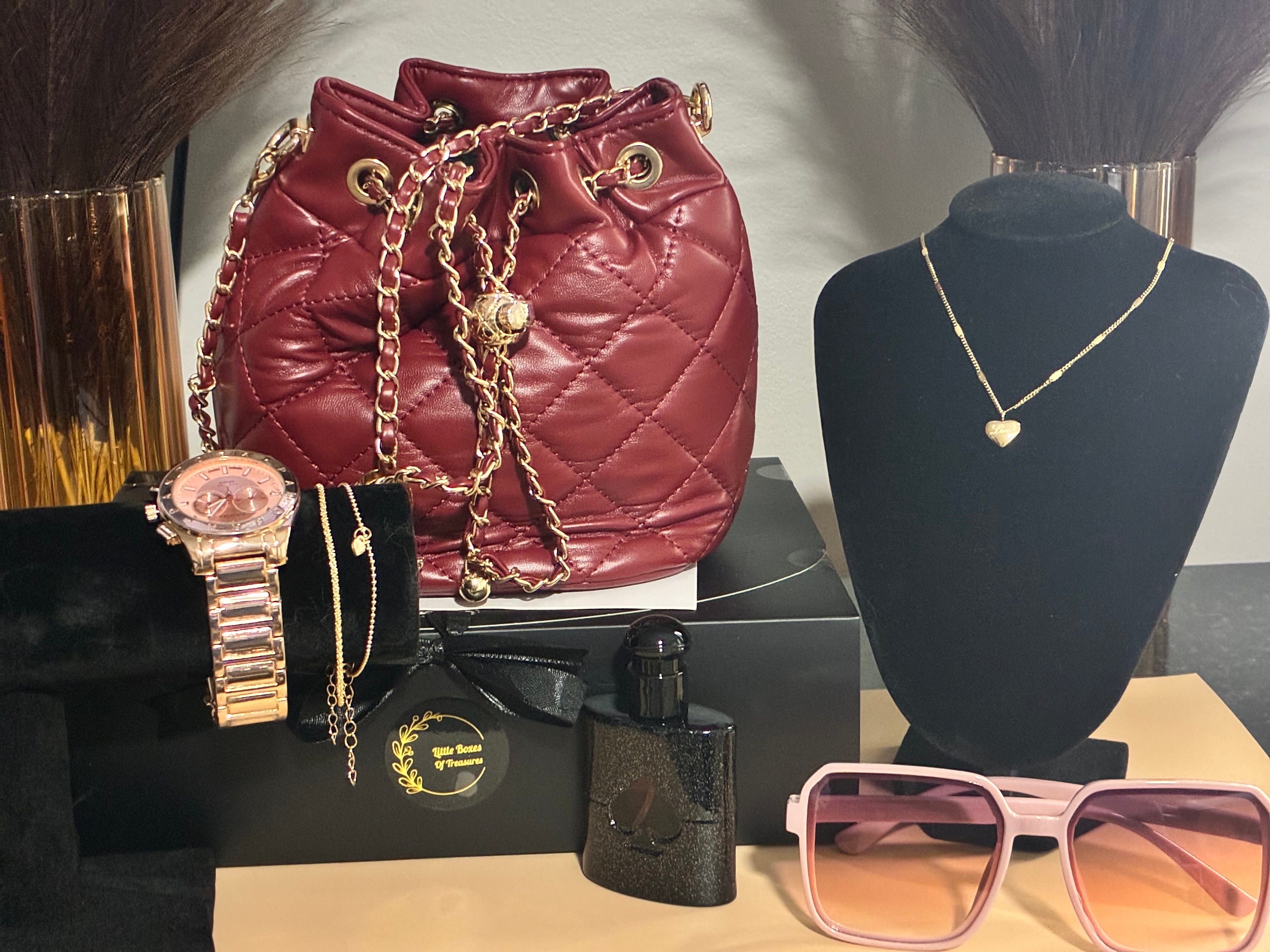 Maroon quilted handbag with gold chain, pink sunglasses, black watch, and perfume bottle on a table.