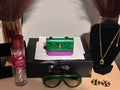 Green and purple handbag on a black stand with a bottle, sunglasses, and jewelry on a table.