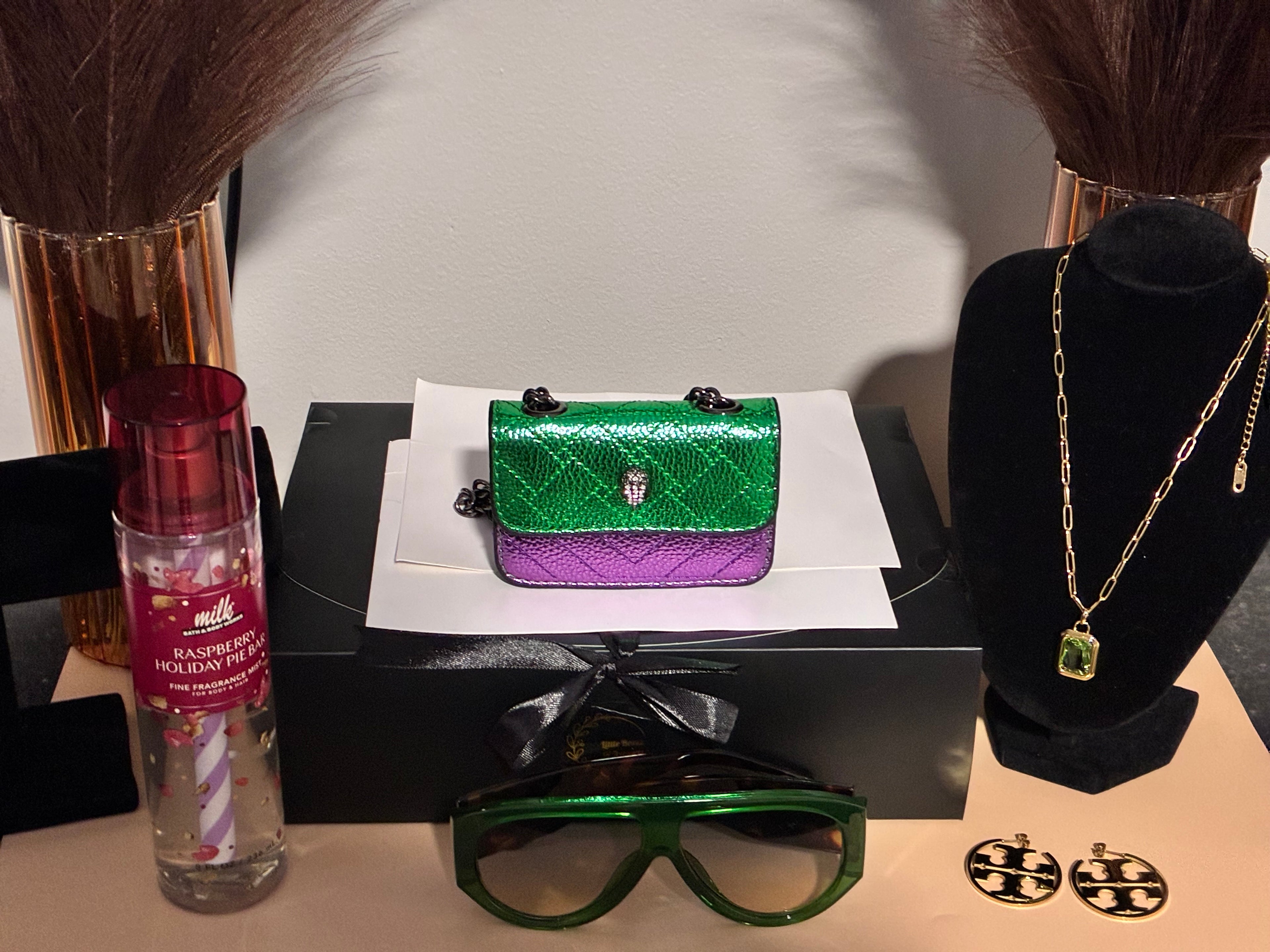 Green and purple handbag on a black stand with a bottle, sunglasses, and jewelry on a table.
